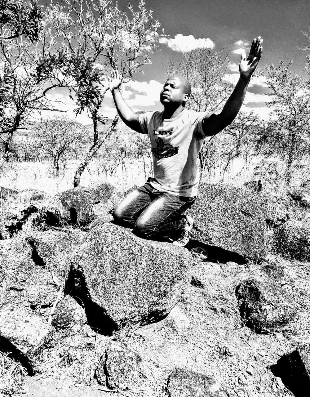 Prophet Matimba Nyandane kneeling on rocks outdoors with his hands lifted in prayer